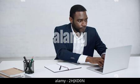 serious african american businessman typing on laptop near notebook on grey Stock Photo