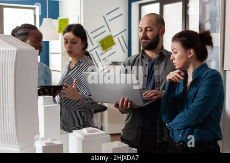 Team of professional architects meeting at office analyzing blueprints on laptop computer and digital tablet. Architectural engineers doing teamwork next to maquette building model project. Stock Photo