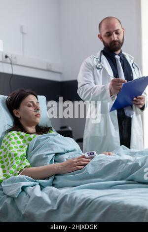 Woman with low oxygen saturation wearing nasal cannula and pulse oximeter recovering after pulmonary surgery in hospital bed. Medic doing rounds writing on patient chart after consult in private ward. Stock Photo