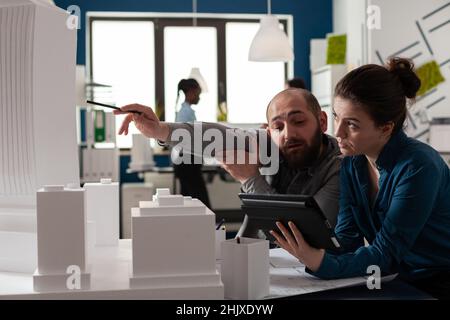 Architectural engineer pointing pen at skyscraper foam model collaborating with colleague engineer holding tablet with blueprints. Architects working together discussing building design. Stock Photo