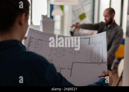 Over shoulder view of architect holding blueprints in architectural office doing teamwork with collegue engineer in the background. Closeup of construction plans of modern business floor plan. Stock Photo