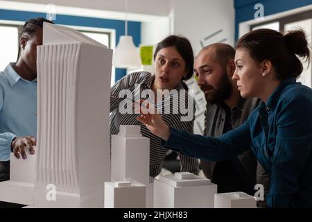 Professional architect with working partners pointing at skyscraper foam scale model in architectural office. Team of engineers collaborating on group project at desk with buildings maquette. Stock Photo