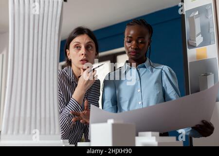 Professional team of two architects inspecting skyscraper white foam model while holding blueprints. Project engineers colleagues looking inspired in front of 3d residential maquette. Stock Photo