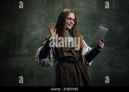 Vintage portrait of young beautiful girl in image of medieval warlike woman using modern gadget isolated over dark background. Comparison of eras Stock Photo