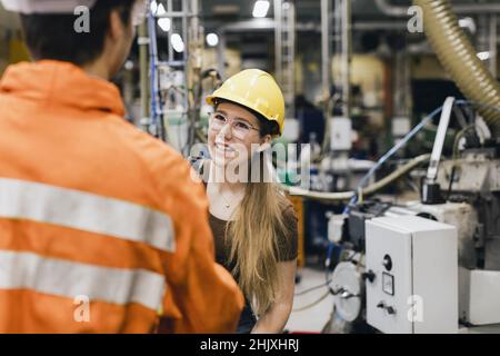 Smiling female manager wearing hardhat talking with male worker in factory Stock Photo