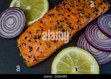 hot smoked salmon steak flavored with black pepper with lemon slices and red onion slices on a slate tray Stock Photo
