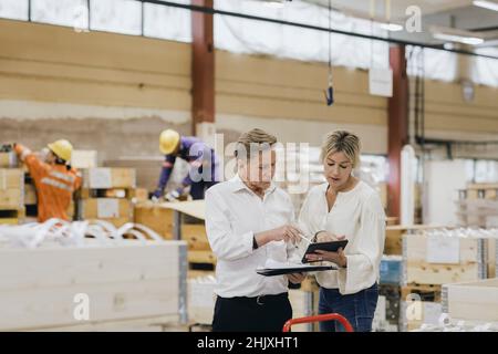 Businesswoman and businessman discussing over digital tablet while workers working in background at warehouse Stock Photo