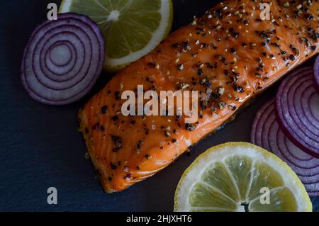 hot smoked salmon steak flavored with black pepper with lemon slices and red onion slices on a slate tray Stock Photo