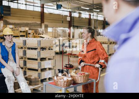 Smiling young female worker with food on push cart talking with colleague in warehouse Stock Photo