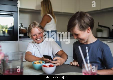 Smiling brothers using smart phone while mother doing kitchen chores in background at home Stock Photo