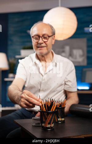 Selctive focus on elderly man illustrator selecting sharp hb pencil to create black and white drawing in home workshop. Older artist choosing professional writing instrument to sketch masterpiece. Stock Photo