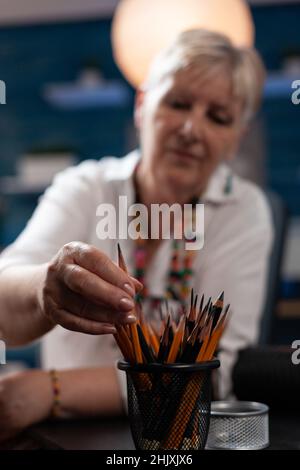 Selective focus on retired woman illustrator selecting hb pencil to create black and white drawing in home workshop. Older artist choosing graphite writing instrument to sketch masterpiece. Stock Photo