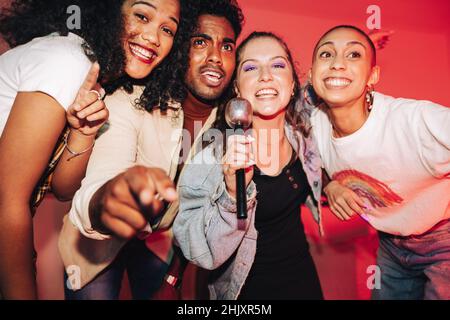 Happy friends singing into a microphone on karaoke night. Group of cheerful friends singing their favourite song at a house party. Multicultural frien Stock Photo