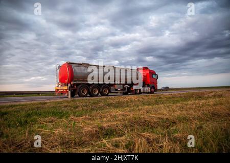 New red truck with a tanker truck transports gasoline against the background of the cloudy sky in the evening. Dangerous goods transport industry. Cop Stock Photo