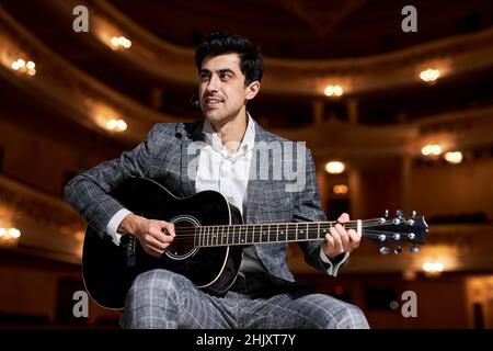 caucasian european Man in elegant classic suit plays and sings while standing on stage on 12-string acoustic guitar, performance. Lighted background o Stock Photo