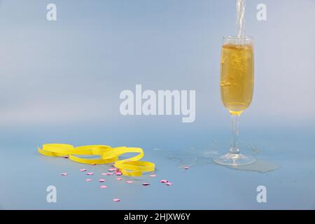 glass of sparkling wine is poured, pink confetti and yellow streamers on the table, blue background, happy new year, studio shot without people Stock Photo