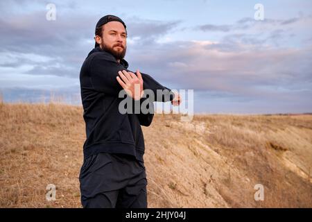 bearded guy in black sportswear stretching arms warming up muscles before running, jogging. caucasian male training outdoors alone, looking confident Stock Photo