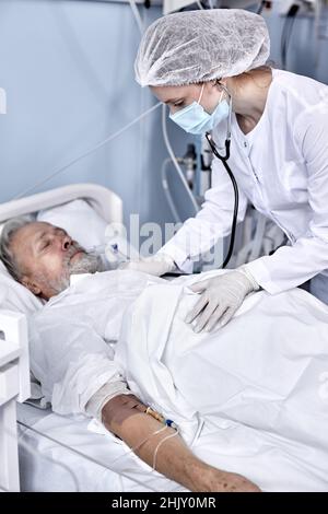 Senior Caucasian Male Patient receiving consultation from doctor using stethoscope for heartbeat exam. Confident Female Nurse consulting retired man w Stock Photo