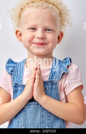 Close up of adorable happy small girl isolated on grey studio background hold hands together, dream, feel grateful, smiling little child, pray thankin Stock Photo
