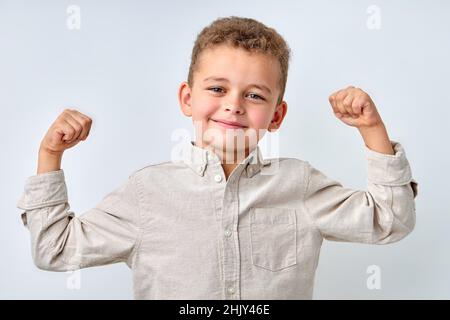 Handsome confident child boy in casual shirt smiling happily and raising clenched fists, tensing muscles, feeling strong and full of energy after ate Stock Photo