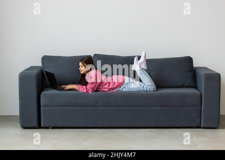 Girl engaged on laptop while lying the sofa in room at home. Concept education is process teaching at school. Teenager studying the computer. Copy spa Stock Photo