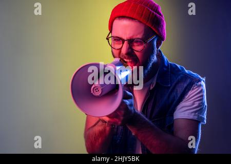 Excited hipster guy screaming loud using megaphone, making announcement, presentation, protesting, wearing beanie hat and denim vest. Indoor studio shot isolated on colorful neon light background. Stock Photo