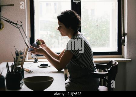 Mature craftswoman painting cup with cleaning sponge while sitting on chair Stock Photo