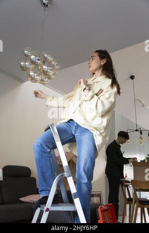 Young woman installing chandelier while relocating at home Stock Photo