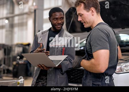 Handsome diverse mechanics in uniform examining car, using laptop while working in auto service. black and caucasian Auto mechanics checking car,servi Stock Photo