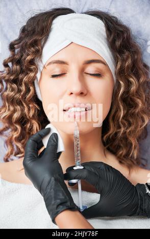 Close up portrait of attractive young woman with curly hair lying and beautician doing injection in lip with special preparation. Concept of process procedure lip augmentation in professional salon. Stock Photo