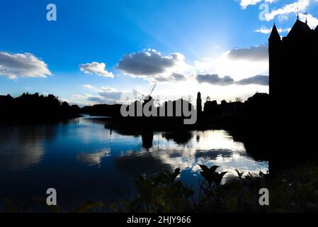 A landscape silhouette of a building and windmill during sunset and the sky is reflected on the water. Stock Photo