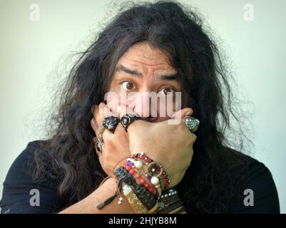 Surprised funny brunette Caucasian young man with big brown eyes and long curly hair covers his mouth with his beringed hands and looks at viewer. Stock Photo