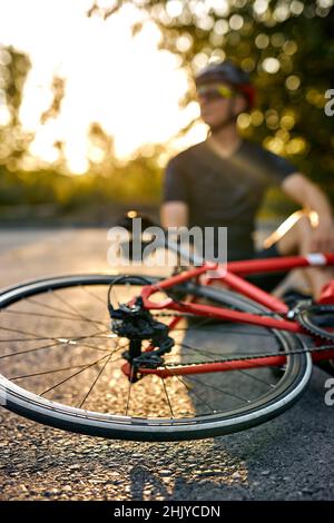 Sporty guy biker in sportive clothes and helmet sit on road near bicycle, having rest. Male resting and relaxing after training on bicycle in nature. Stock Photo