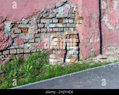An old brick wall with peeling off plaster stands along an asphalt path, green grass nearby, selective focus Stock Photo