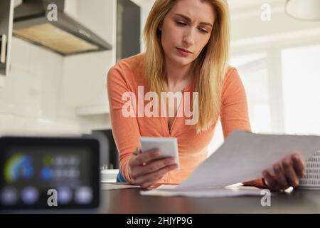 Smart Energy Meter In Kitchen Measuring Electricity And Gas Use With Woman Looking At Bills With Calculator Stock Photo