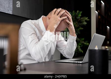 Tired or upset mature businessman sitting at desk with laptop and wrapping head in hands because of problems or overload. Work failure concept. High quality photo Stock Photo