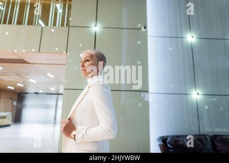 side view of blonde senior businesswoman in white blazer in lobby of modern hotel Stock Photo