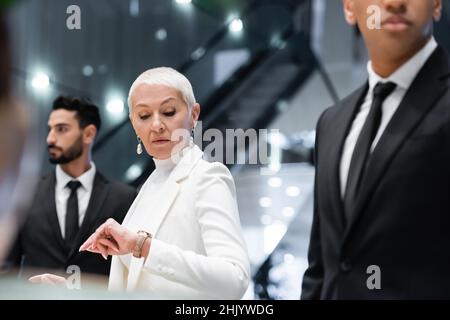 mature businesswoman checking time on wristwatch near interracial bodyguards in hotel