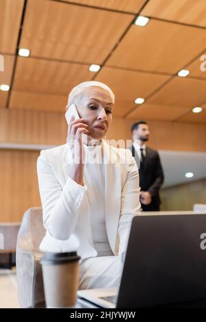 senior business lady talking on smartphone near blurred laptop and bi-racial bodyguard on background Stock Photo