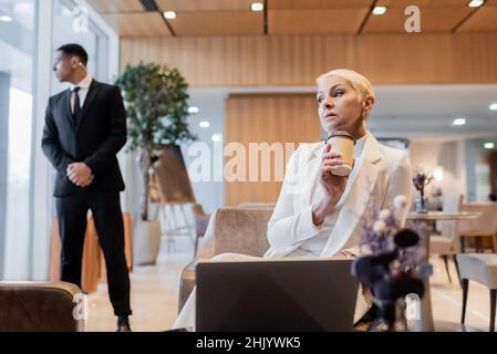 stylish senior businesswoman with coffee to go sitting near laptop and blurred african bodyguard in hotel lounge Stock Photo