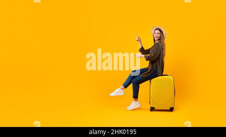 Female Tourist Pointing Fingers Aside Sitting On Suitcase, Yellow Background Stock Photo