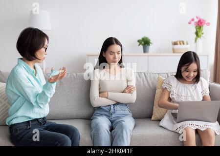 Offended young Asian woman having fight with her mature mother about little girl's gadget addiction at home Stock Photo