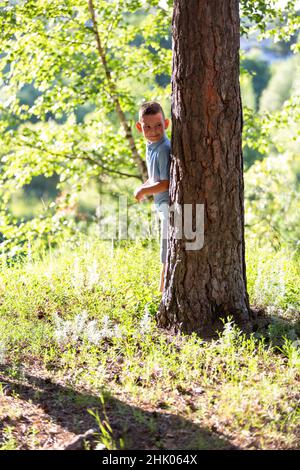 The boy looks out from behind a tree, hides, plays hide-and-seek Stock Photo