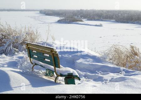 A bench in snowdrifts on a steep bank of a river against the backdrop of a distant forest. Stock Photo