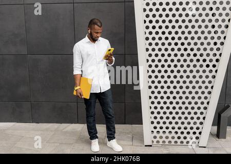 Concentrated stylish business guy wearing casual outfit, using smartphone, chatting with colleague. Handsome serious male freelancer standing outside Stock Photo