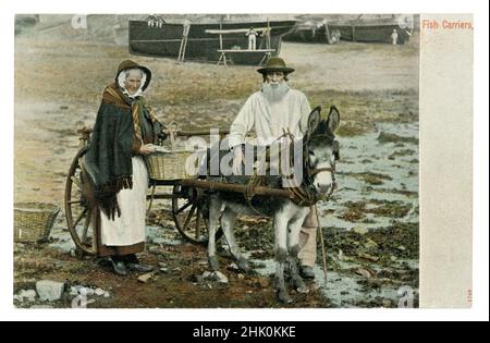 Original Edwardian era tinted greetings souvenir postcard of Cornish Fish Carriers, fishwife, with donkey cart on the waterfront, Newlyn harbour, Cornwall, U.K. The man is a local called Billy Renfree and his fishwife is filling up her basket with fish. She wears a traditional shawl and apron known as ‘towsers’, dated to 1906 from a posted postcard. Stock Photo