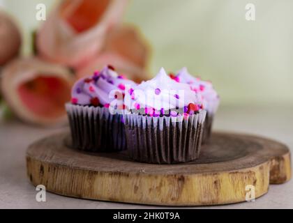 Three chocolate valentine's day cupcakes on a wooden slab and tulips blurred in the background.  Light green background. Stock Photo