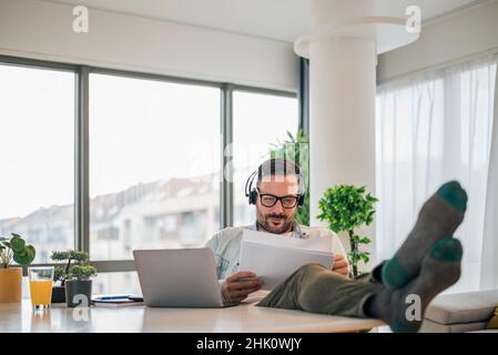 Thoughtful dedicated focused pensive businessman entrepreneur with feet on the table working with documents and laptop computer making a online video Stock Photo