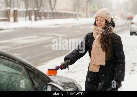 Young woman standing in snowfall and cleaning her car from snow. She is freezing and needs to get to work. Stock Photo