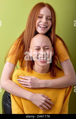 Two diverse european red-haired and bald ladies models hugging isolated over green background, portrait of young females in casual yellow shirts havin Stock Photo